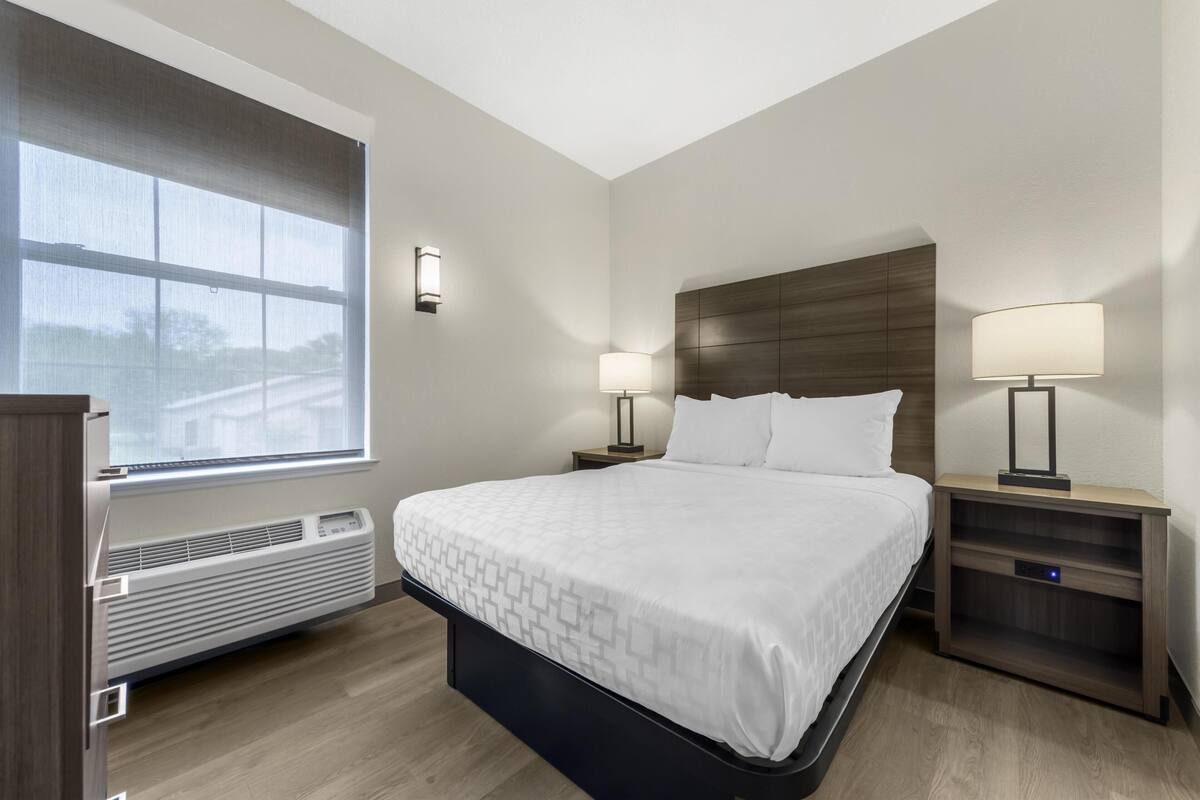 A hotel room with a bed, two bedside tables with lamps, a window, and a built-in heater.