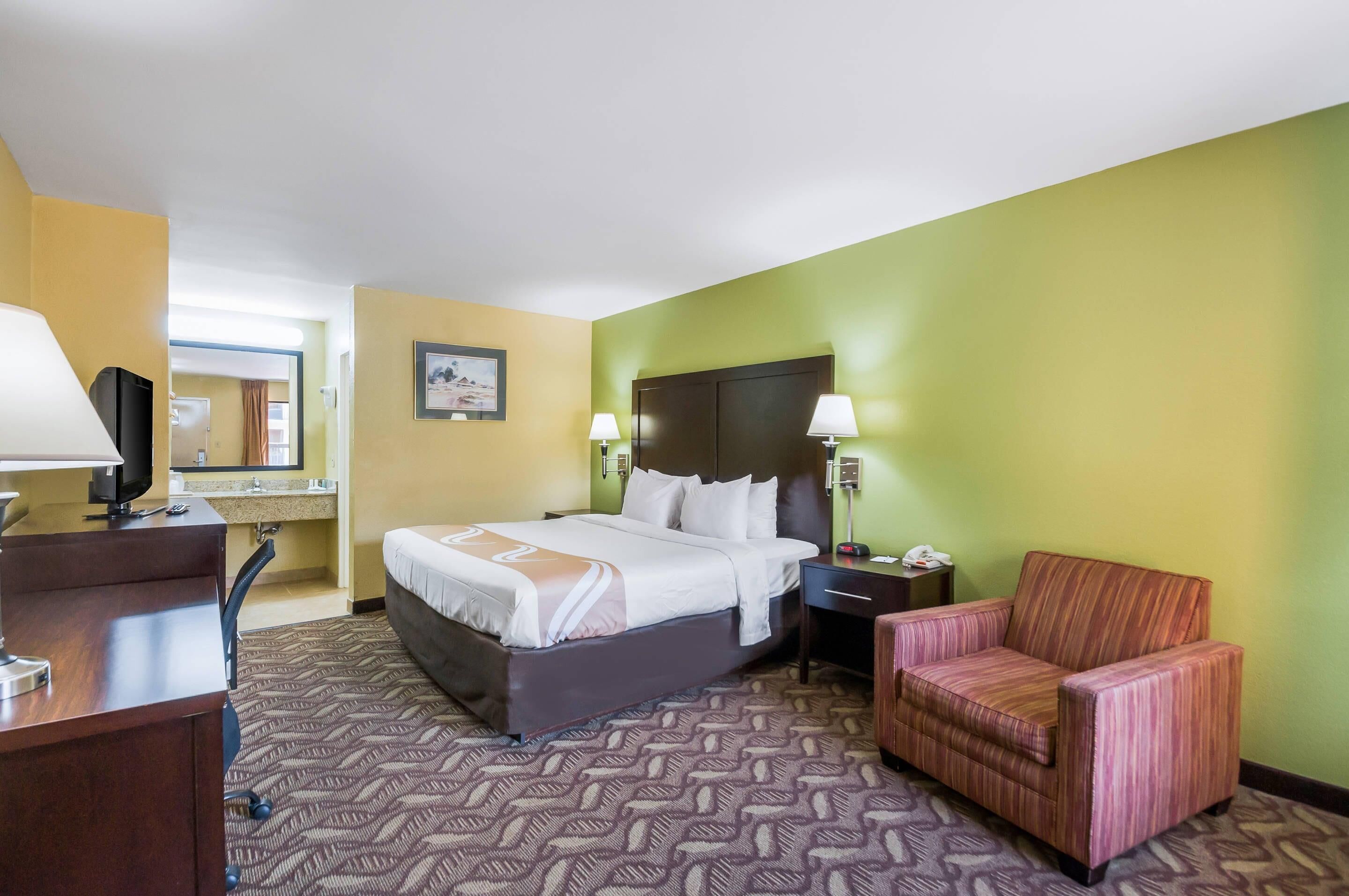 A hotel room with a bed, desk, chair, and a nightstand with a lamp.