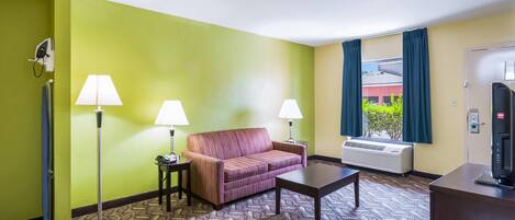 A hotel room with a green wall, a patterned carpet, a sofa, two lamps, a TV, and an air conditioning unit.