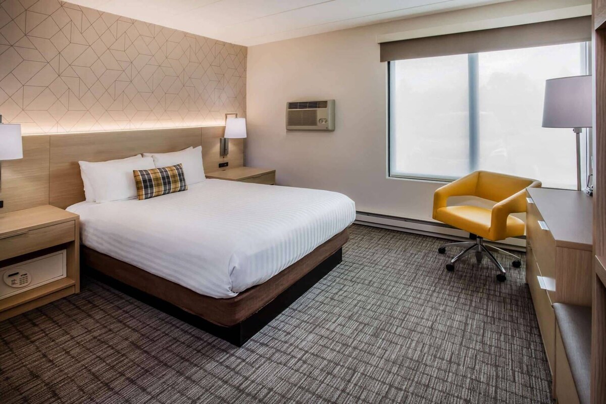 A hotel room with a bed, bedside tables, a desk, a chair, a window, and an air conditioning unit.