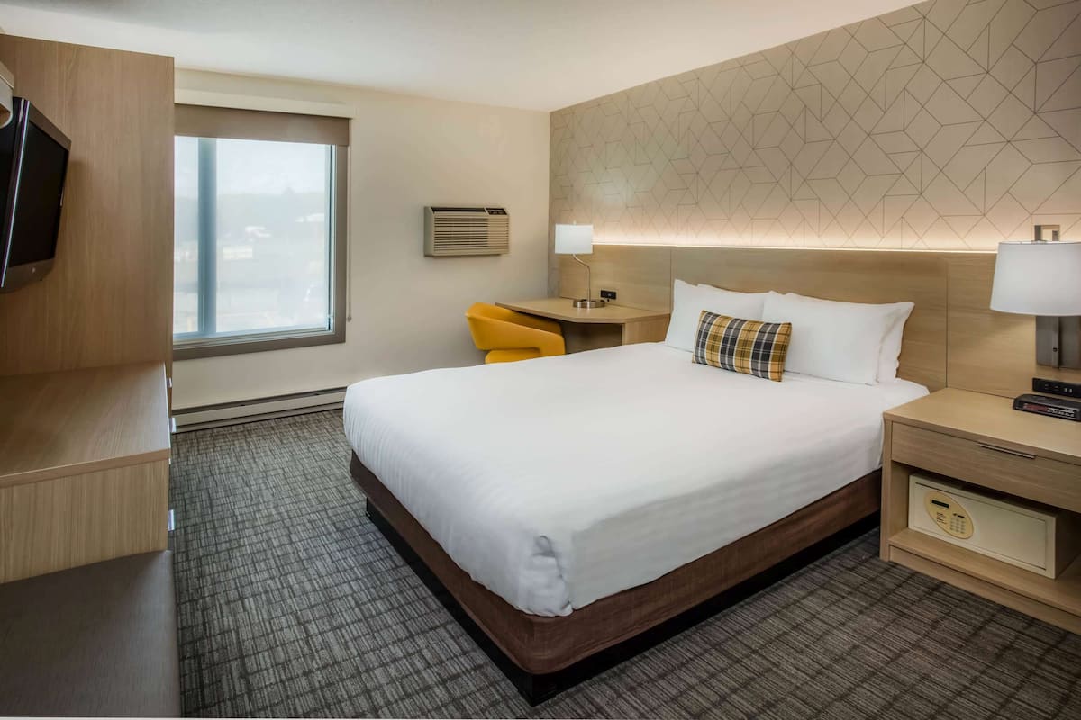 A hotel room with a large bed, a desk with a chair, a TV, an air conditioning unit, and a patterned wall.