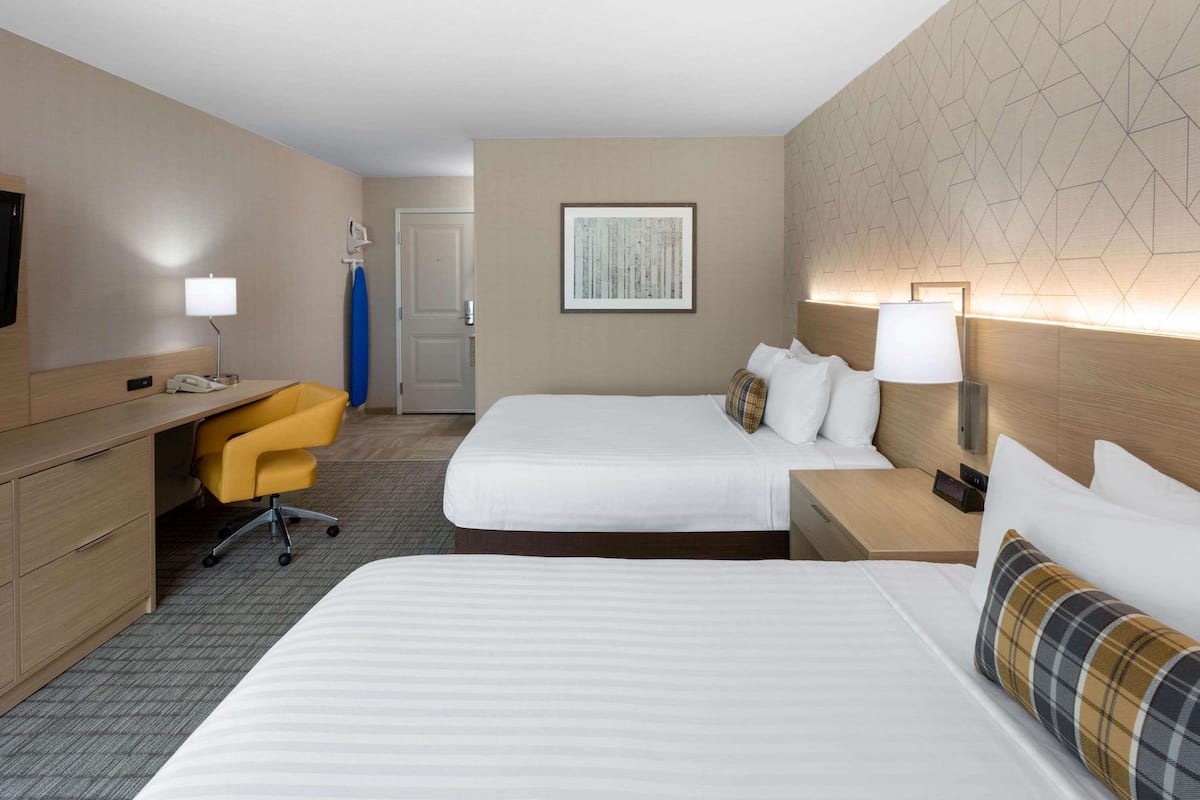 A hotel room with two beds, a desk with a chair, a bedside table with a lamp, and a wall-mounted TV.