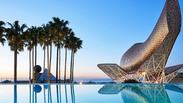 A modern pool area with a unique architectural structure, palm trees, and a clear view of the ocean. - Hotel Arts Barcelona (Barcelona)