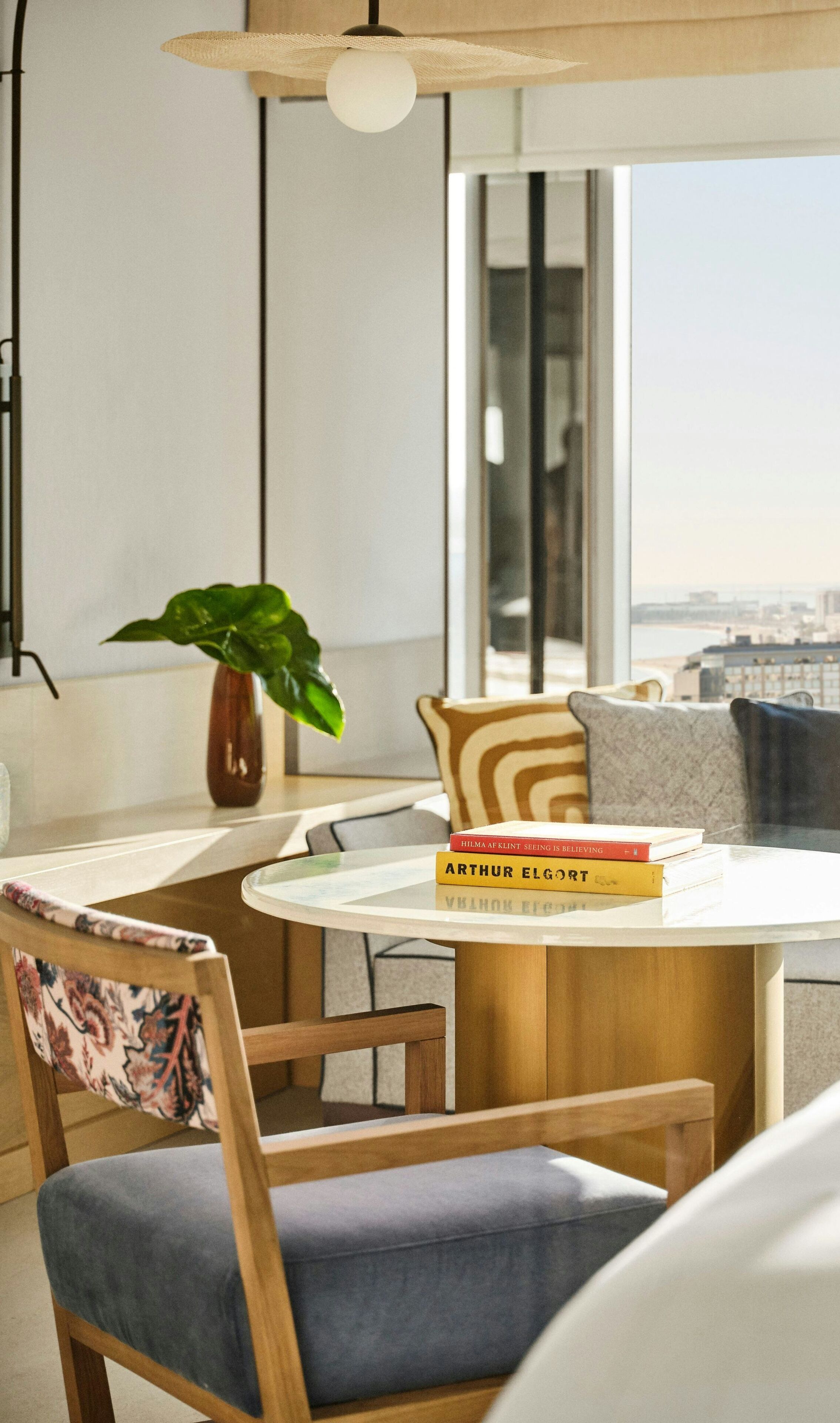 A modern living room with a round table, two chairs, a potted plant, and a view of the city through large windows.