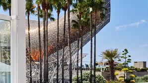 An outdoor seating area with wooden tables and chairs, surrounded by palm trees and a modern building with a unique geometric facade. - Hotel Arts Barcelona (Barcelona)