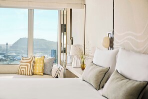 Quarto de hotel moderno com uma janela grande, uma cama com várias almofadas, uma mesa de cabeceira com abajur e um vaso com flores secas.