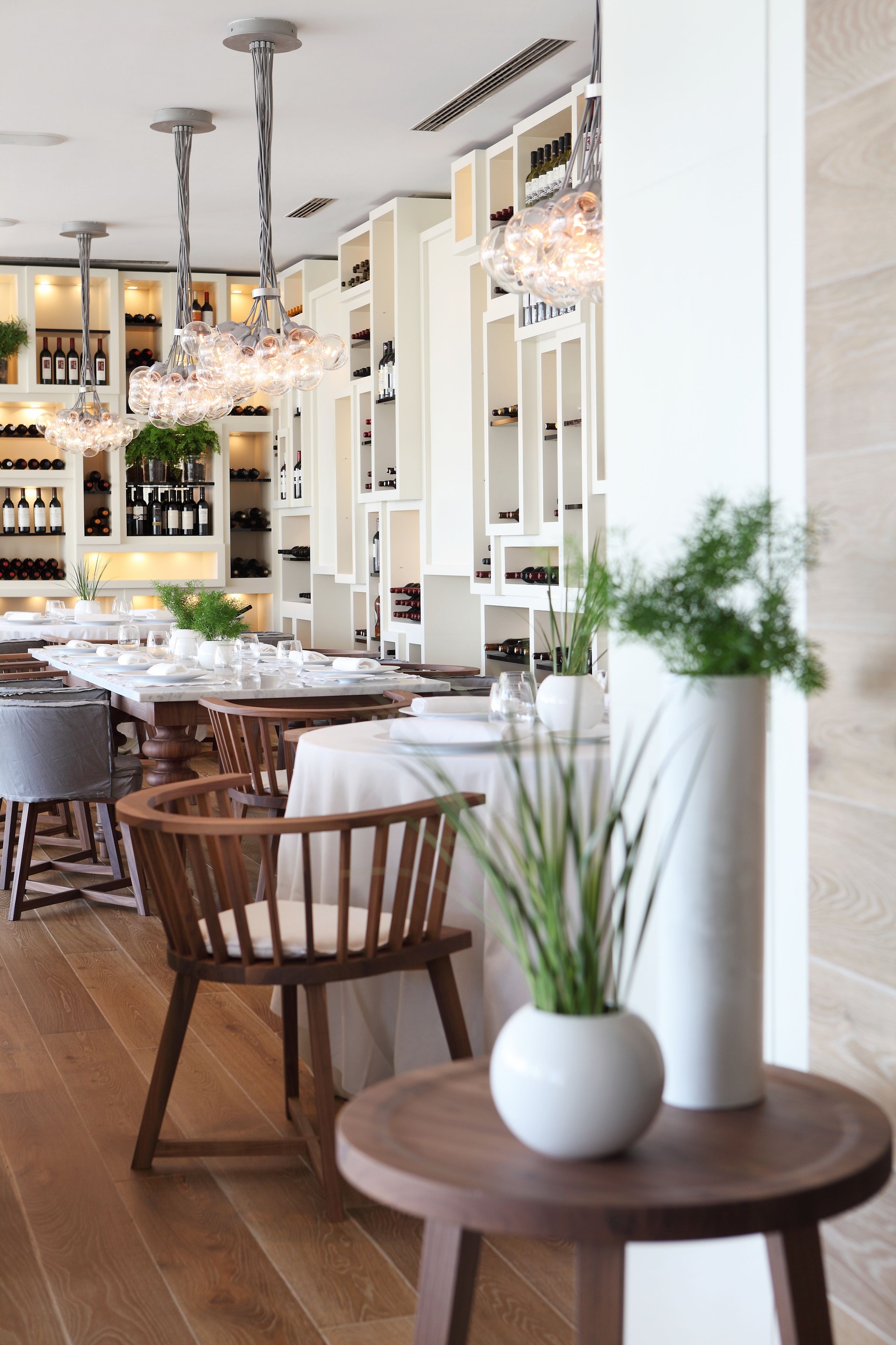 A restaurant with wooden chairs and tables, white shelving with wine bottles, and hanging light fixtures.