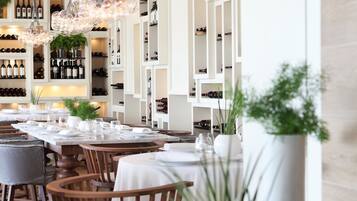 A restaurant with wooden chairs and tables, white shelving with wine bottles, and hanging light fixtures.