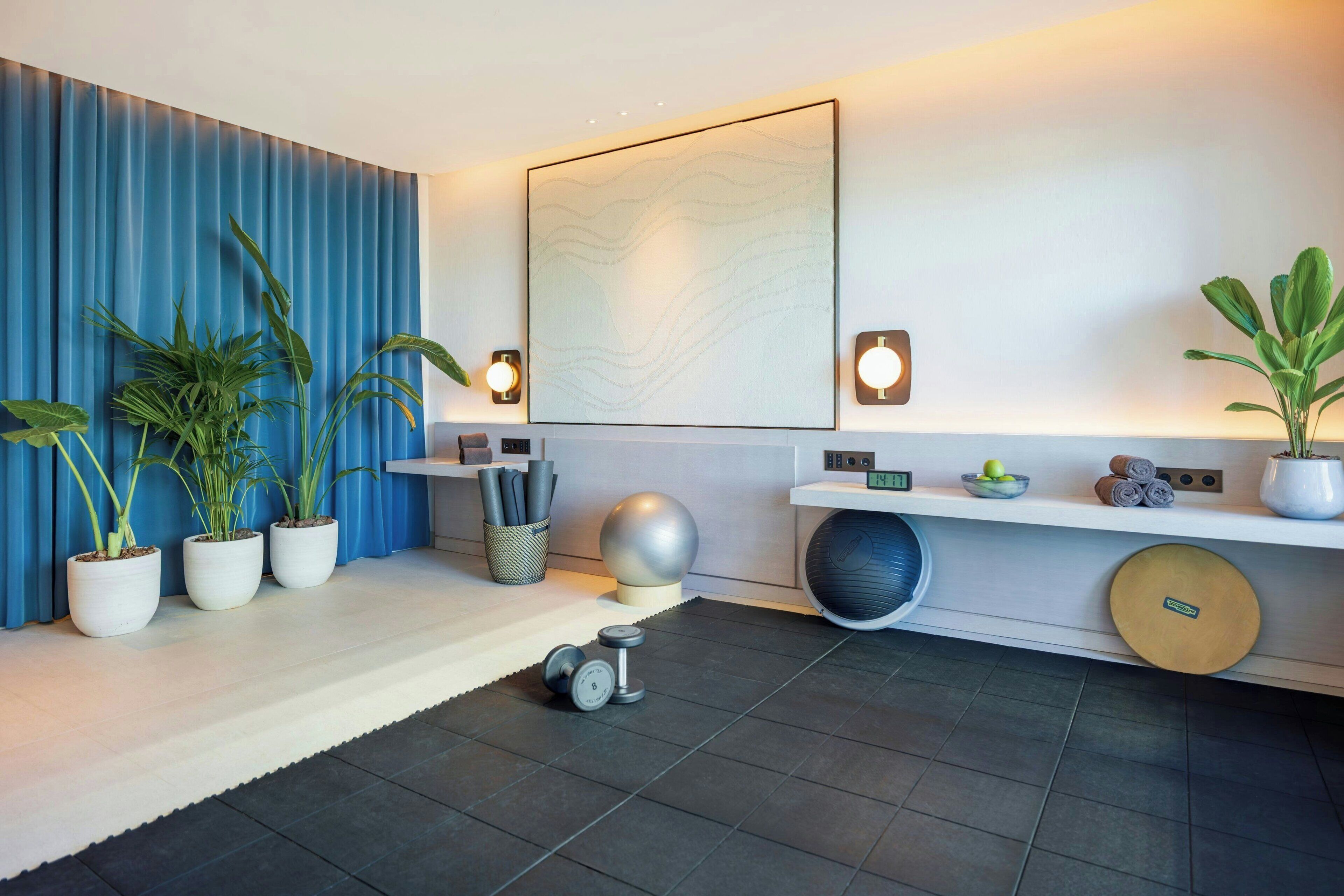 A modern room with a blue curtain, potted plants, a wall-mounted light fixture, a round mirror, and a white console table with a clock.