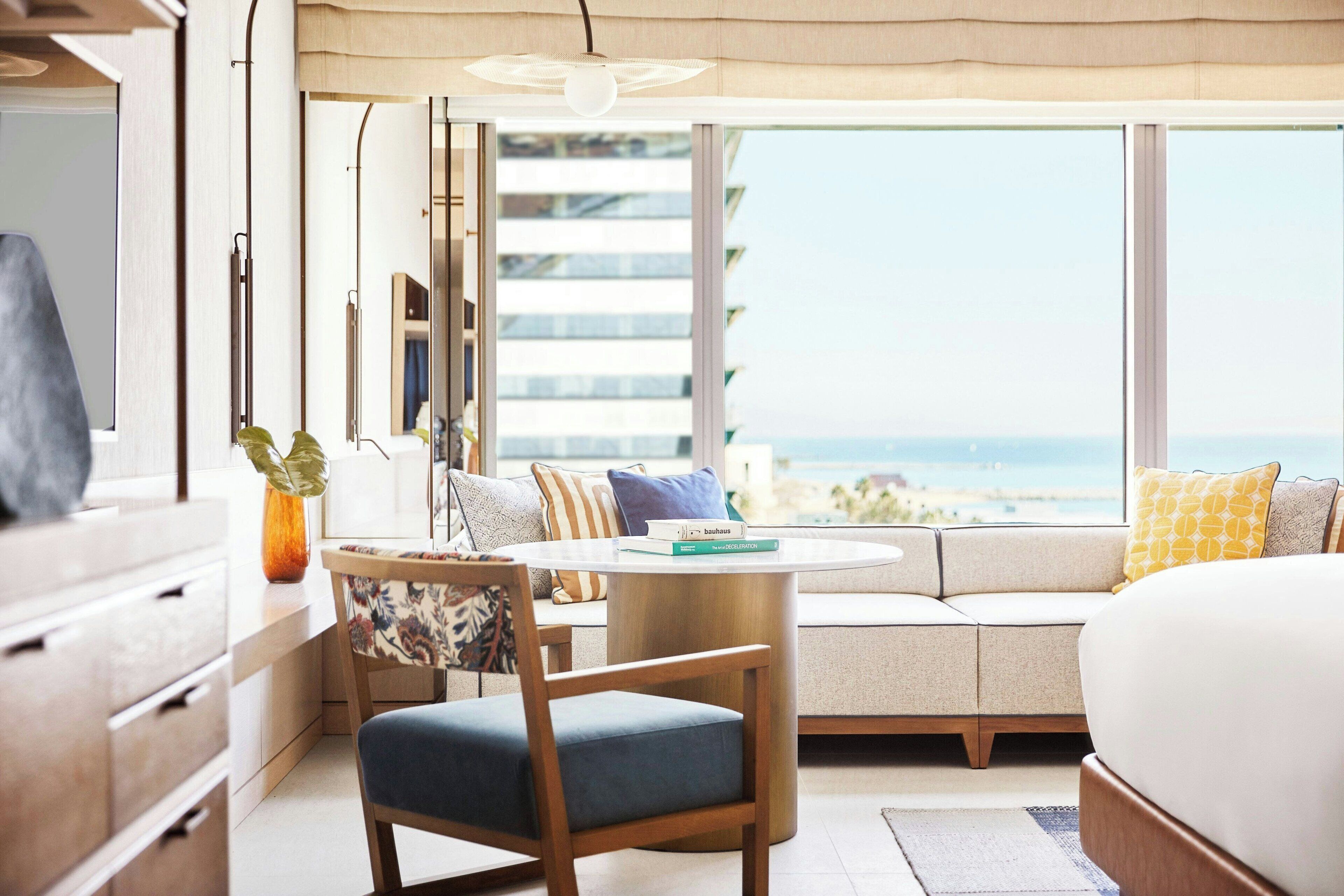 A modern hotel room with a large window overlooking the sea, a round dining table, a sofa, and a bed.