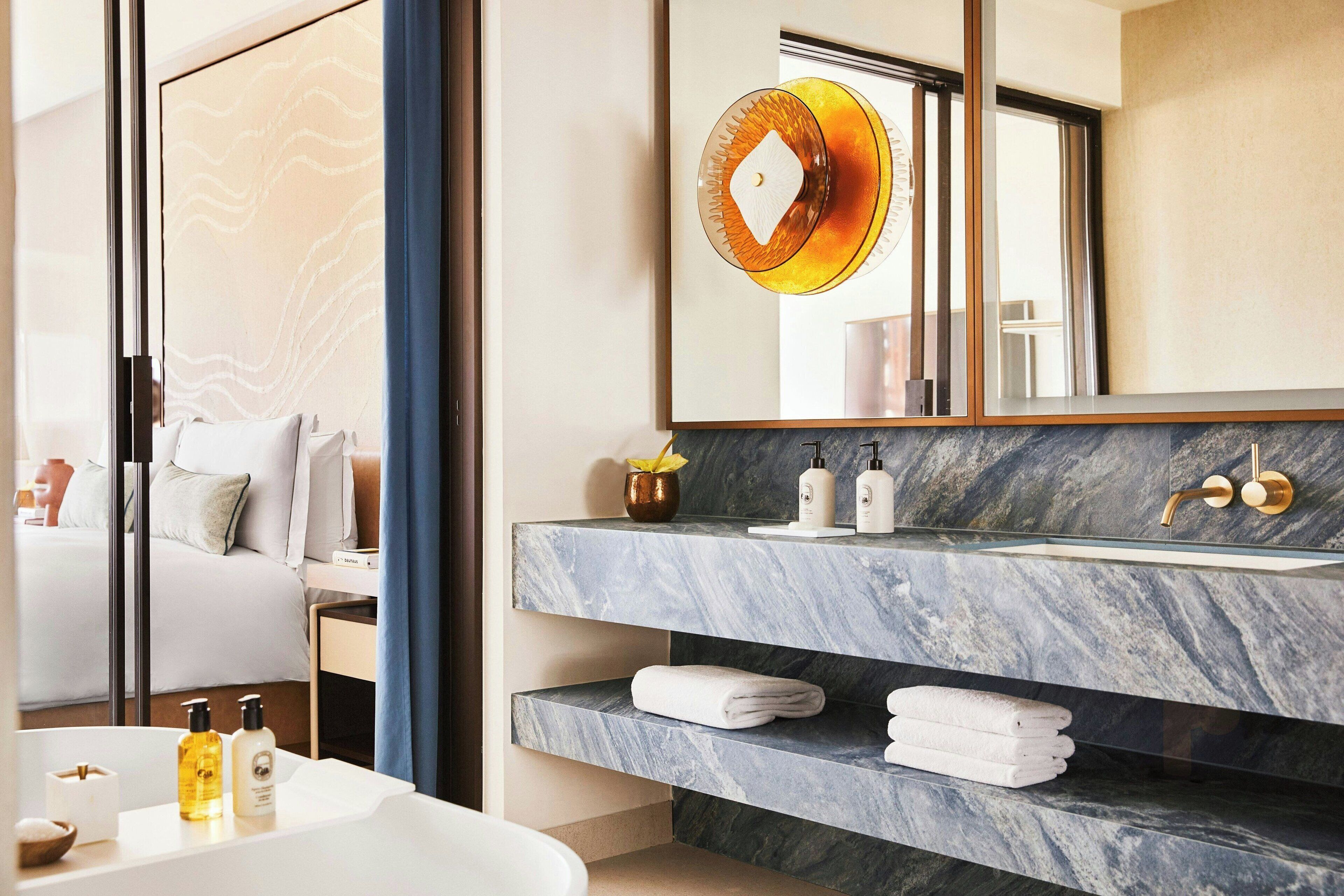 Un baño moderno con tocador de mármol, un espejo grande y una cama visible en una habitación contigua.