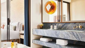 Un baño moderno con tocador de mármol, un espejo grande y una cama visible en una habitación contigua.