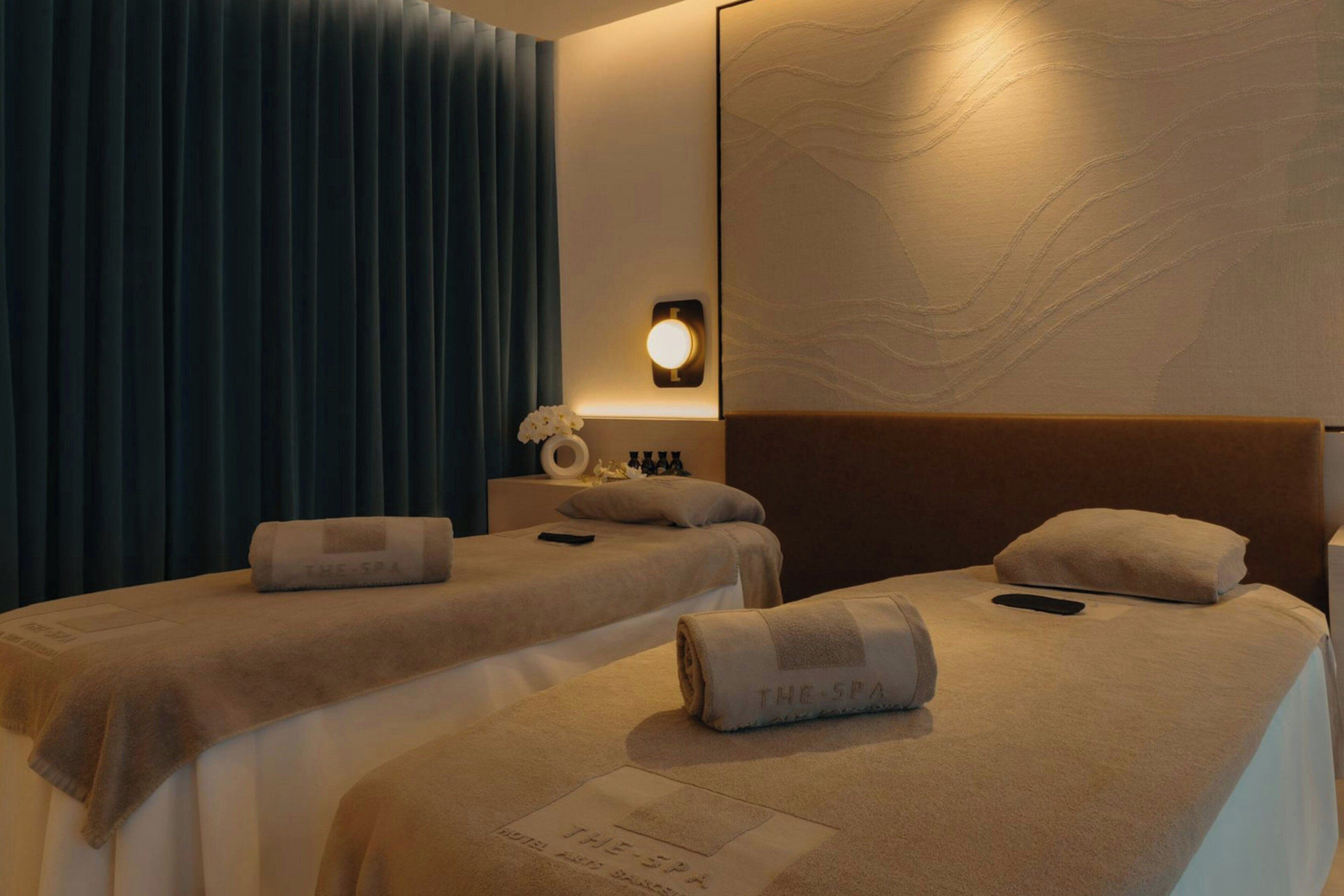 Habitación de hotel con dos camas, cada una con una toalla y un almohadón, una mesita de noche con una lámpara y un espejo grande en la pared.