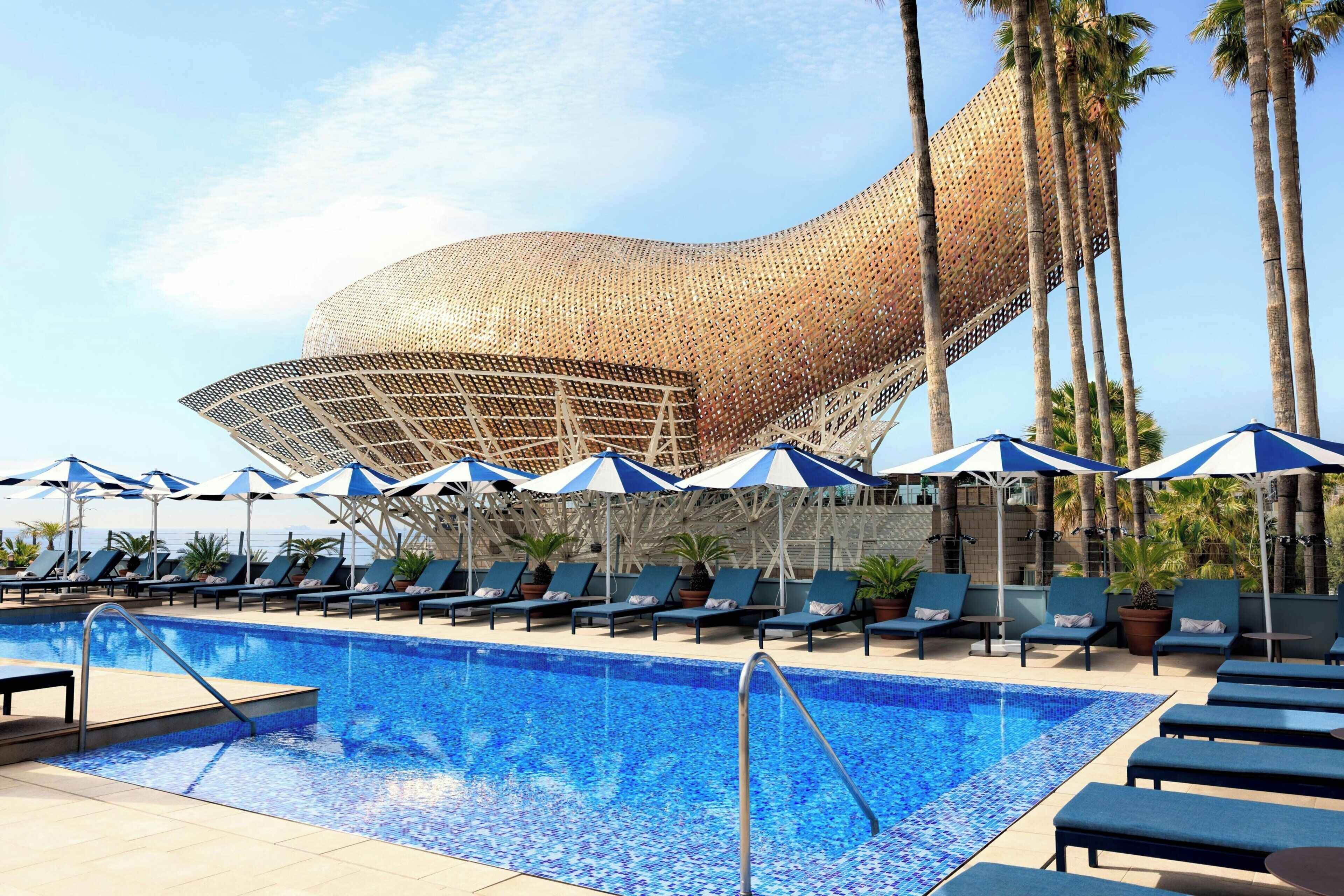 A pool area with lounge chairs and umbrellas, a distinctive building in the background, and palm trees.