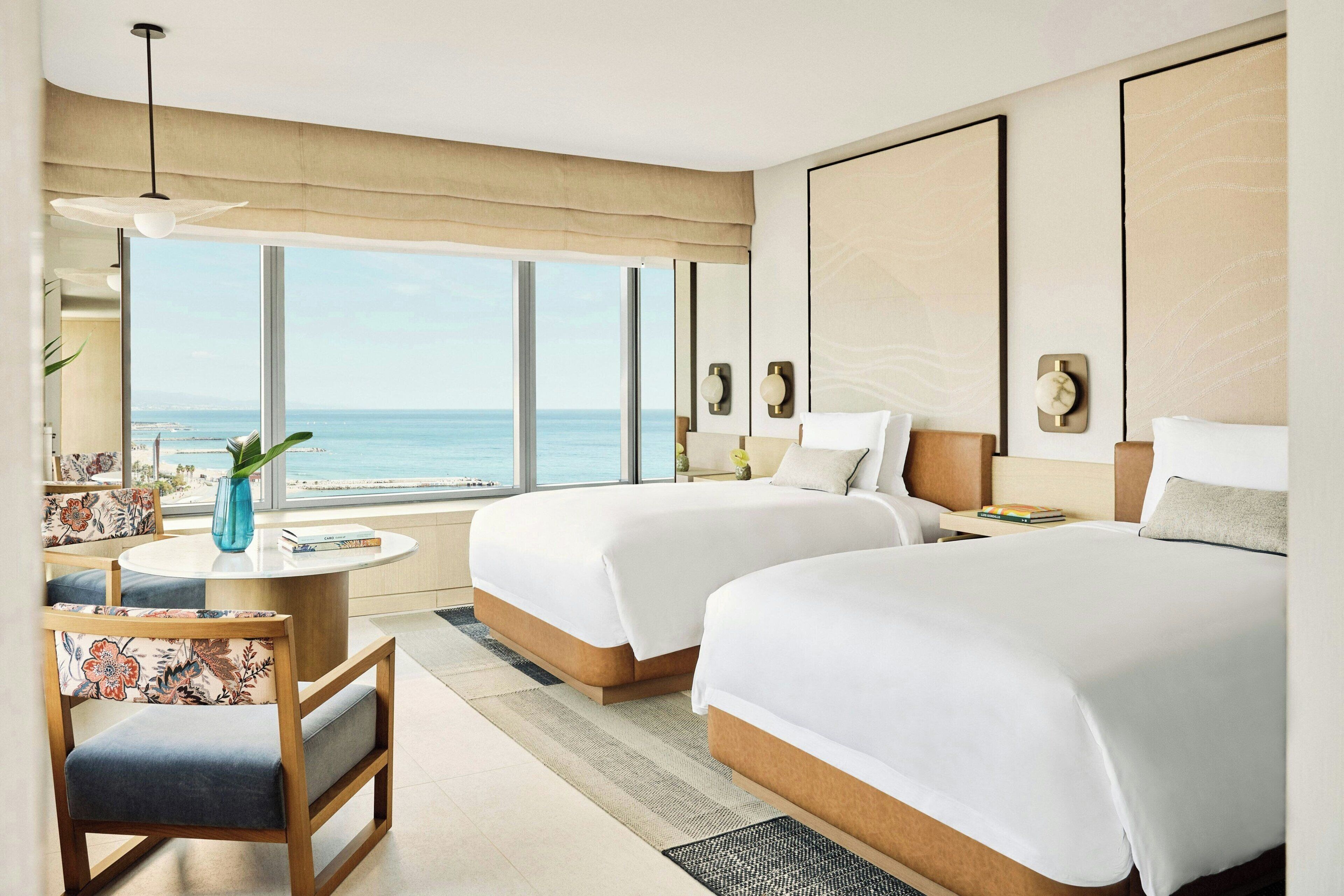 A hotel room with two beds, a round table, a chair, and a large window overlooking the sea.