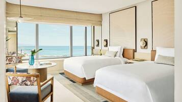 Ein Hotelzimmer mit zwei Betten, einem runden Tisch, einem Sessel und einem großen Fenster mit Meerblick.