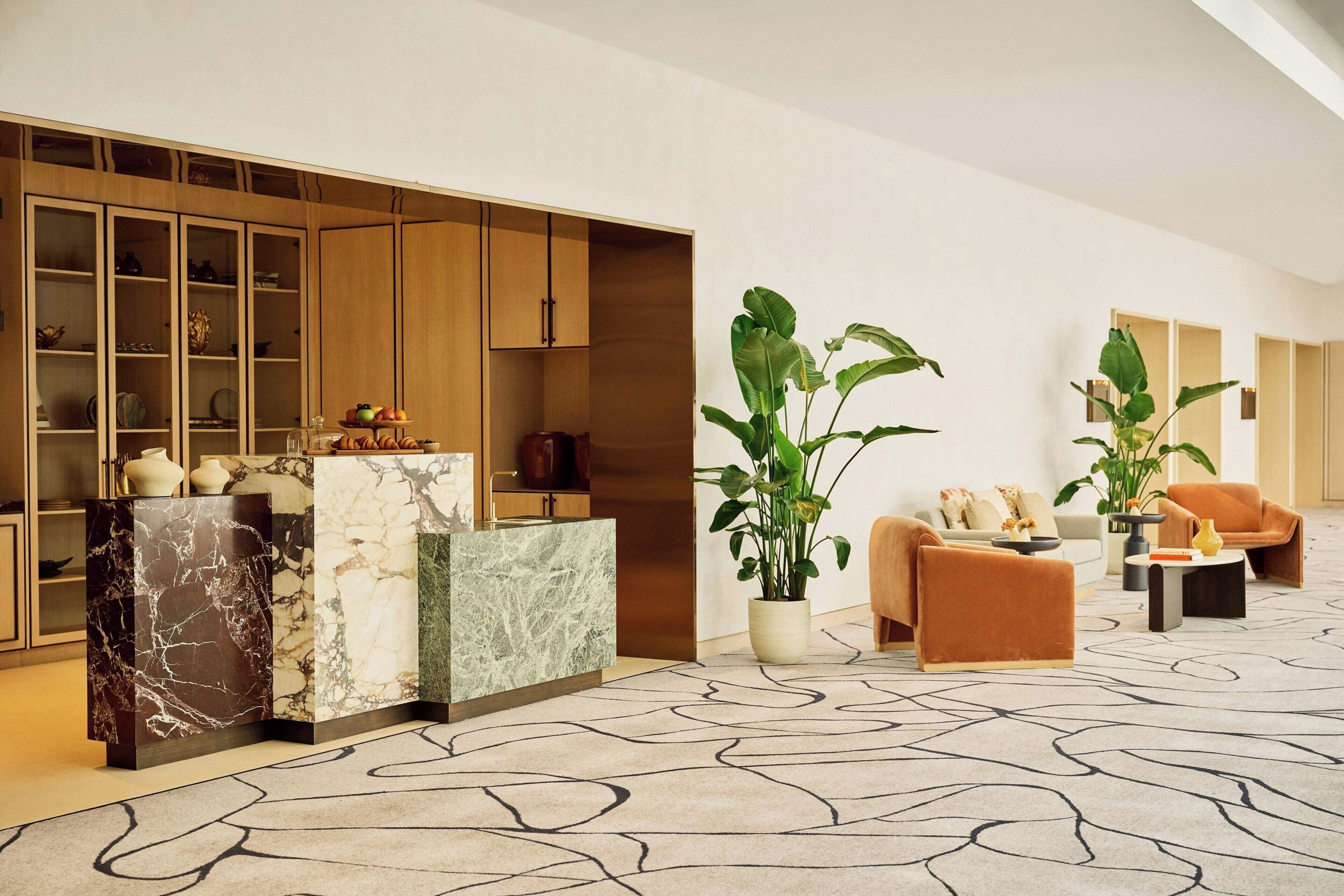 Eine moderne Lobby mit einem Marmor-Empfangstresen, Holzschränken, einer Sitzecke mit Sofa und Sesseln sowie großen Kübelpflanzen.