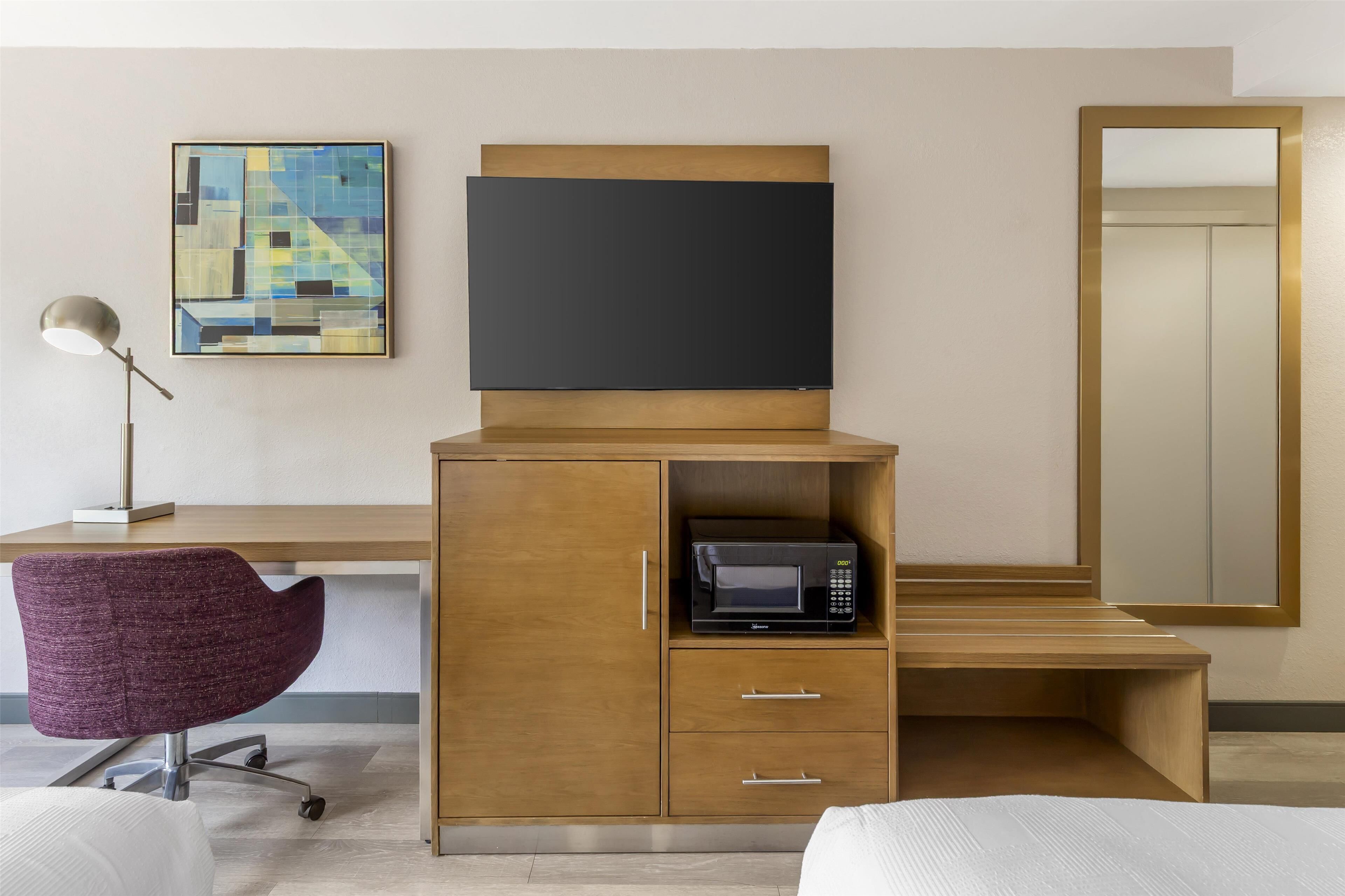 Habitación de hotel con un escritorio de madera, una silla de oficina púrpura, un televisor de pantalla plana, un microondas y un espejo grande.