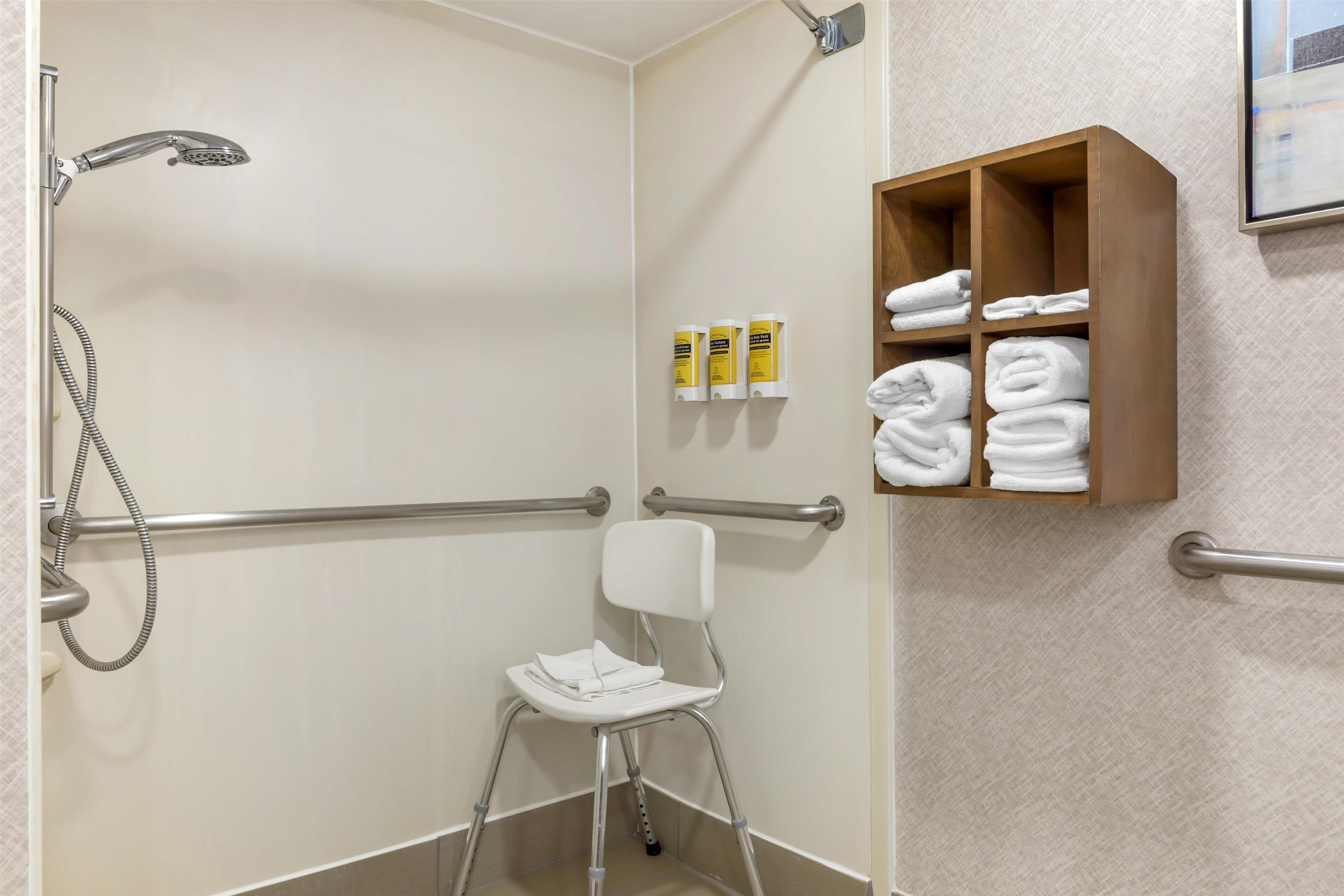 A bathroom with a shower, handrails, a white chair, and a shelf with towels and toiletries.
