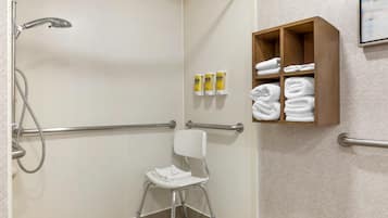 A bathroom with a shower, handrails, a white chair, and a shelf with towels and toiletries.