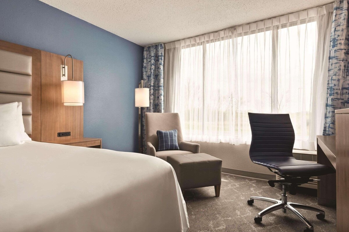 Een hotelkamer met een groot bed, een bureau met stoel, een kleine fauteuil, een lamp en een raam met dunne gordijnen.