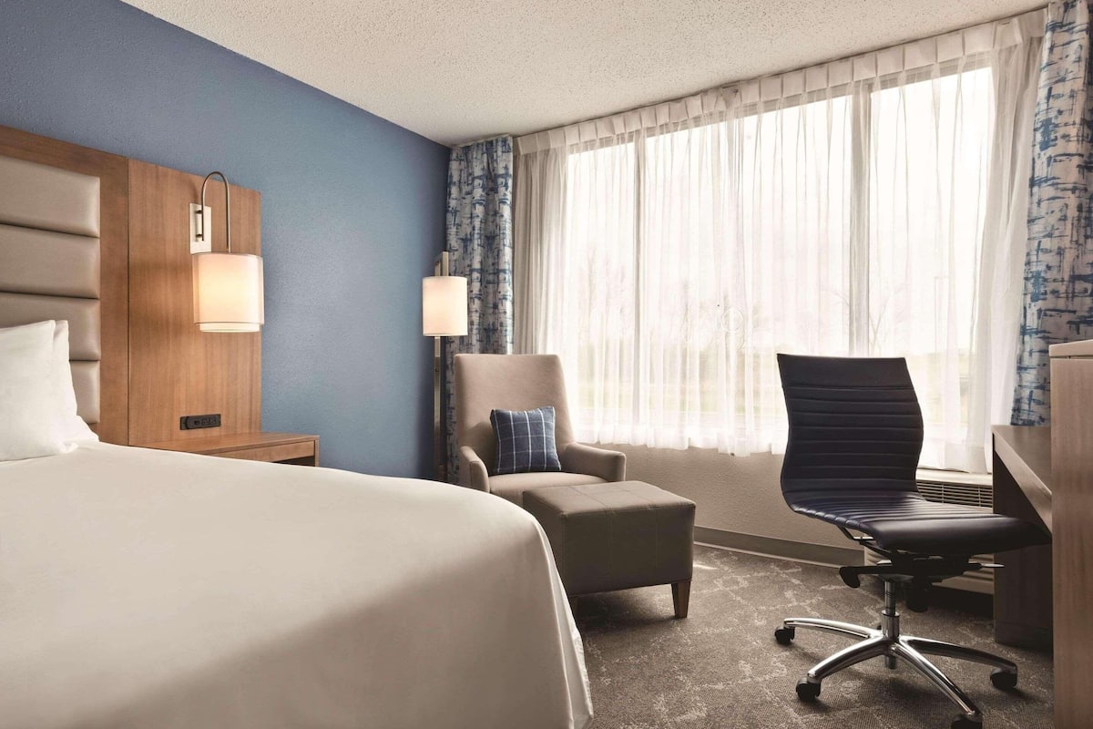 A hotel room with a large bed, a desk with a chair, a small armchair, a lamp, and a window with sheer curtains.