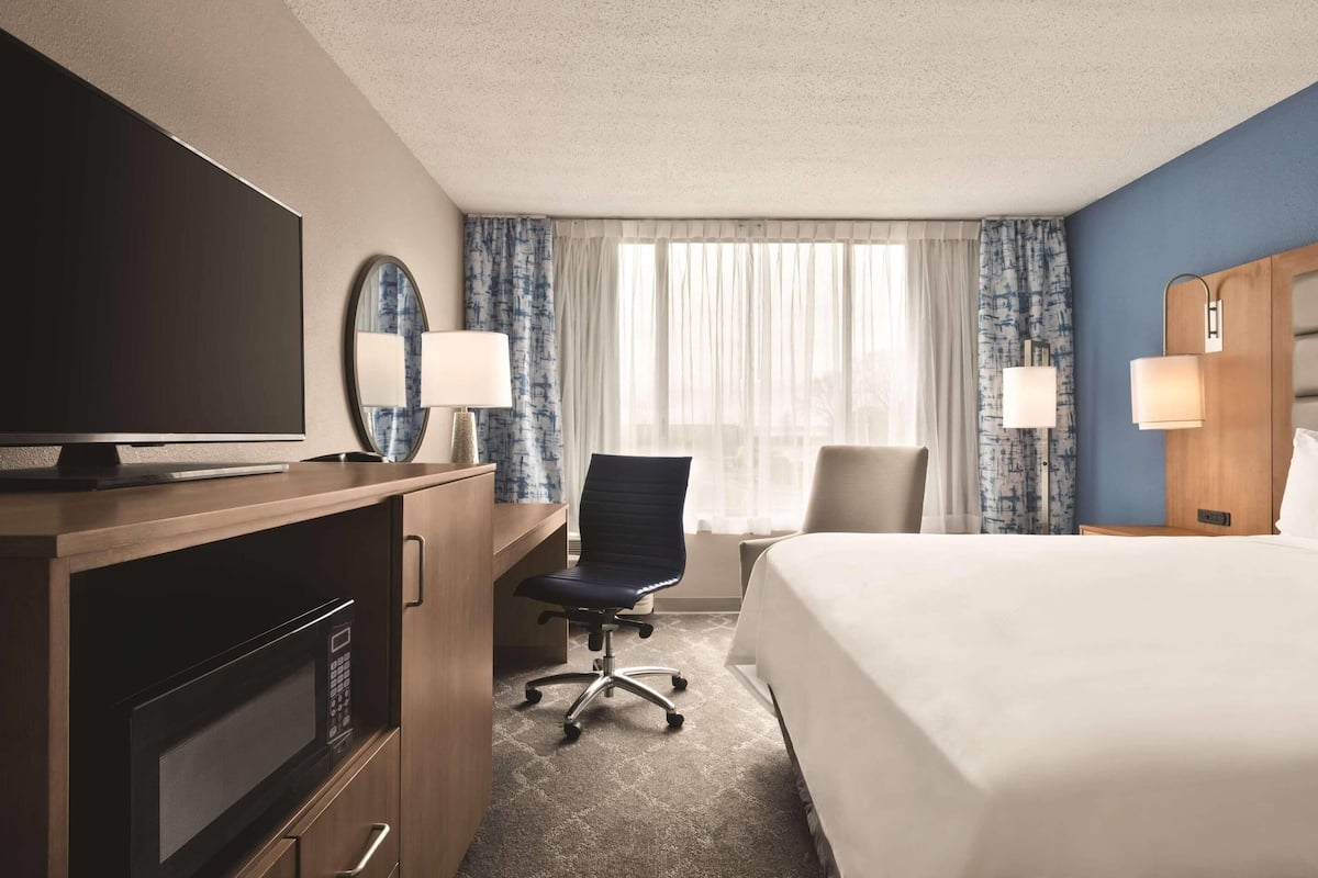 A hotel room with a bed, a desk with a TV, a chair, a desk lamp, and a window with curtains.