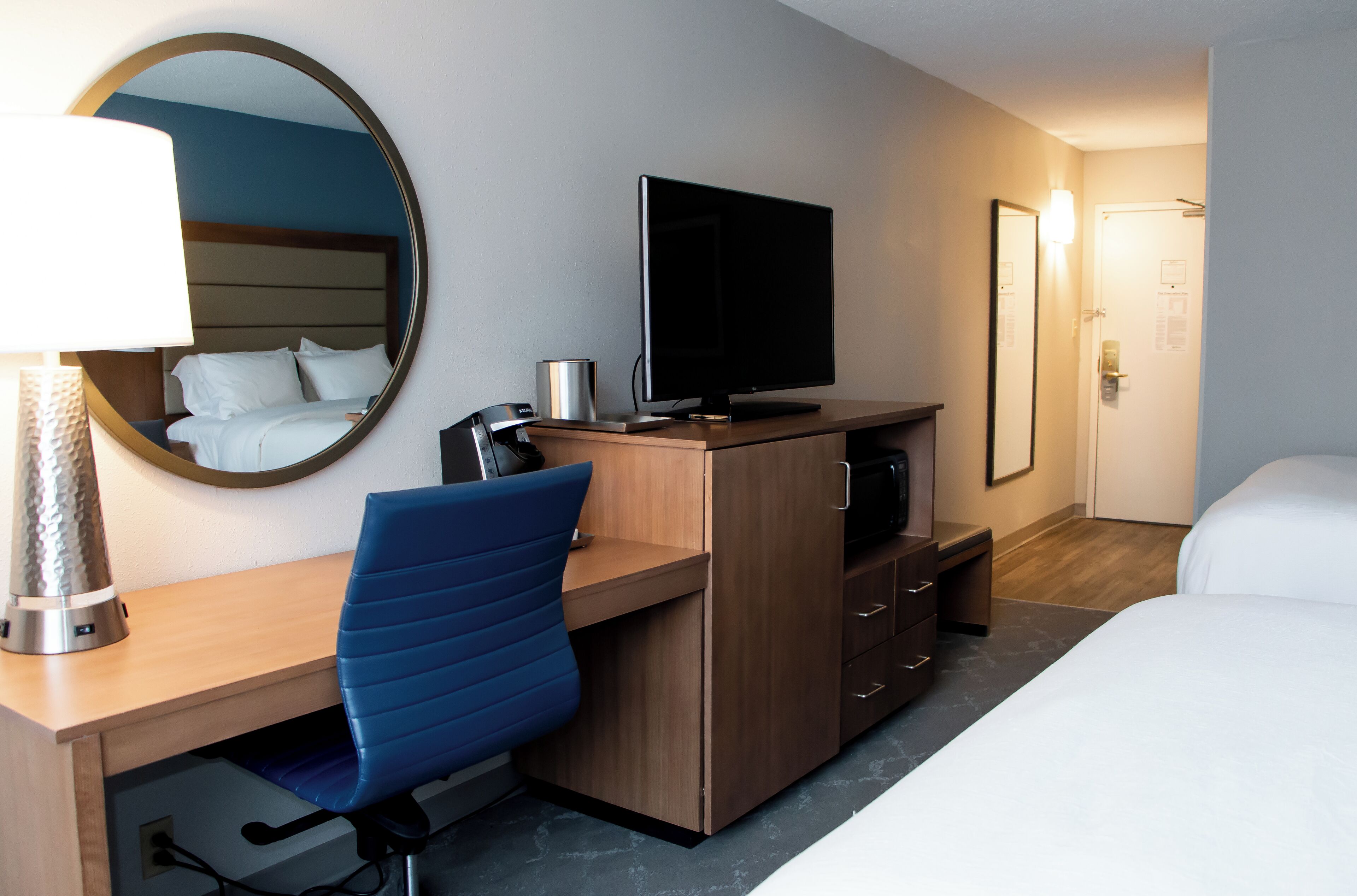 Quarto de hotel com cama, escrivaninha com computador, cadeira, televisão e espelho.