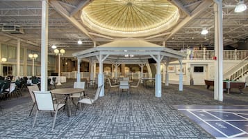 A spacious indoor area with a central open pavilion, multiple tables and chairs, and a shuffleboard court.