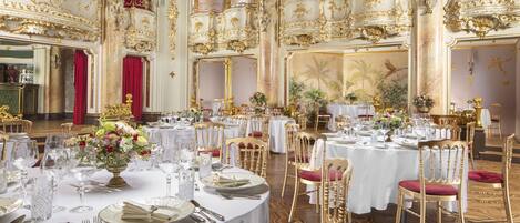 A luxurious banquet hall with ornate gold detailing, red curtains, and elegantly set tables.