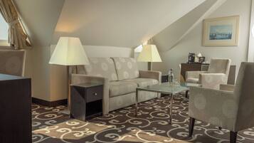 Habitación de hotel con sofá, sillones, mesa de centro de vidrio, lámparas, mesita auxiliar y alfombra estampada.