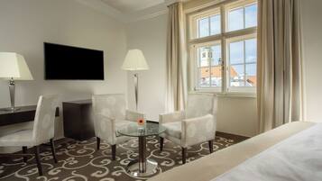 A hotel room with a bed, two armchairs, a small glass table with a vase, a flat-screen TV, and a window with curtains.