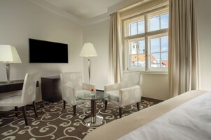 Quarto de hotel com cama, duas poltronas, uma mesinha de vidro com um vaso, televisão de tela plana e janela com cortinas.