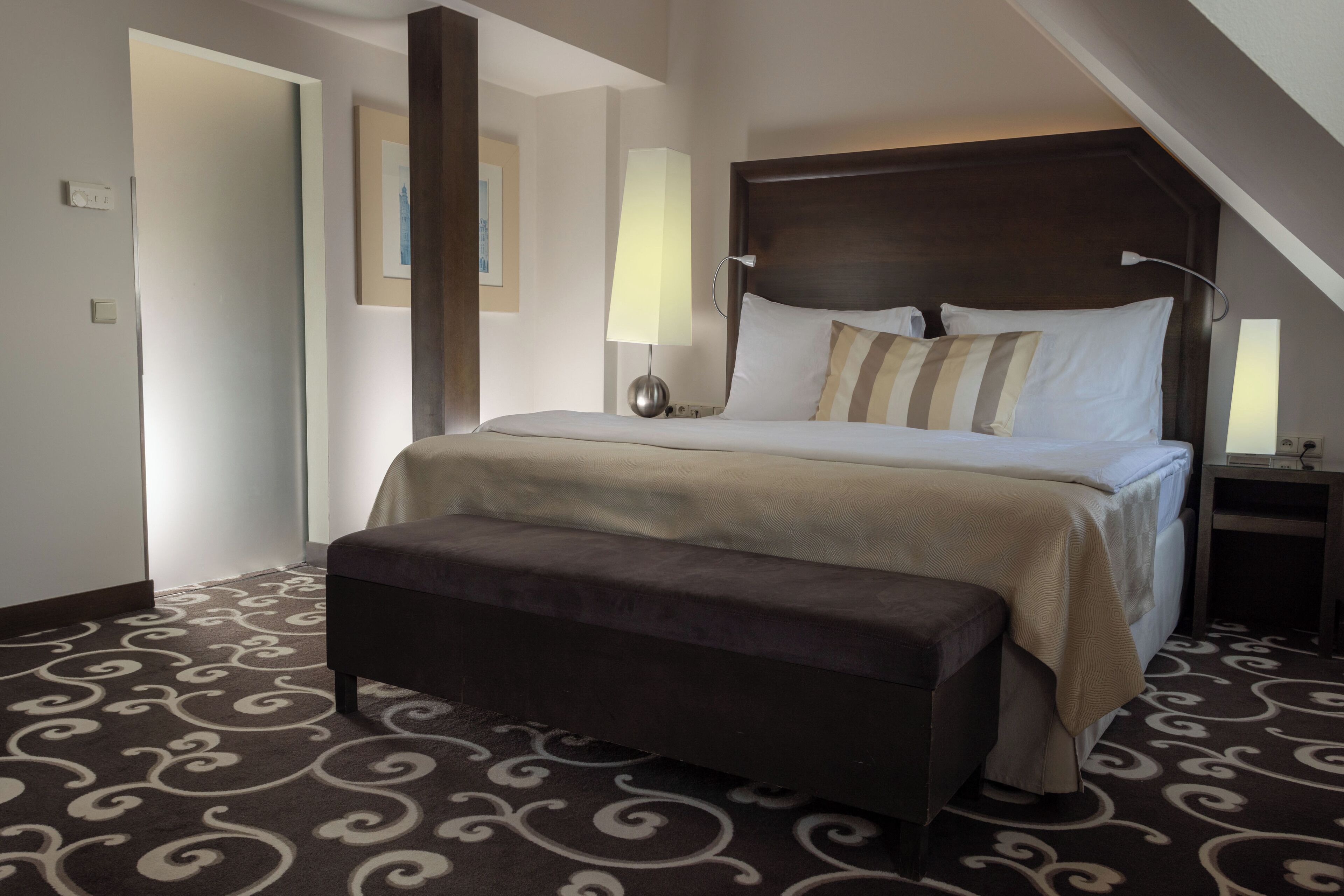 Una habitación de hotel con una cama grande, una mesita de noche con lámpara, un banco y una alfombra estampada.