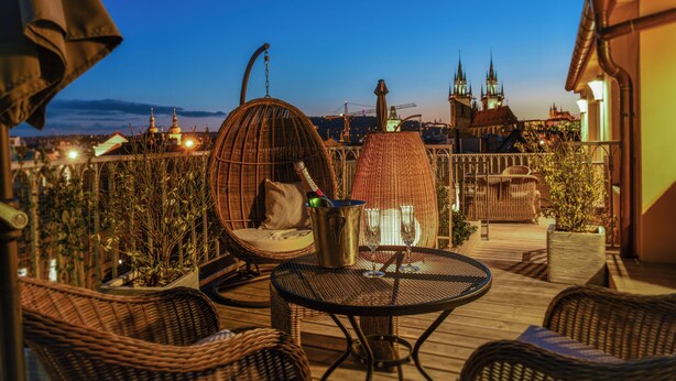 A rooftop terrace with wicker furniture, a metal table, and a view of a cityscape with spires. - Grand Hotel Bohemia (Prague)