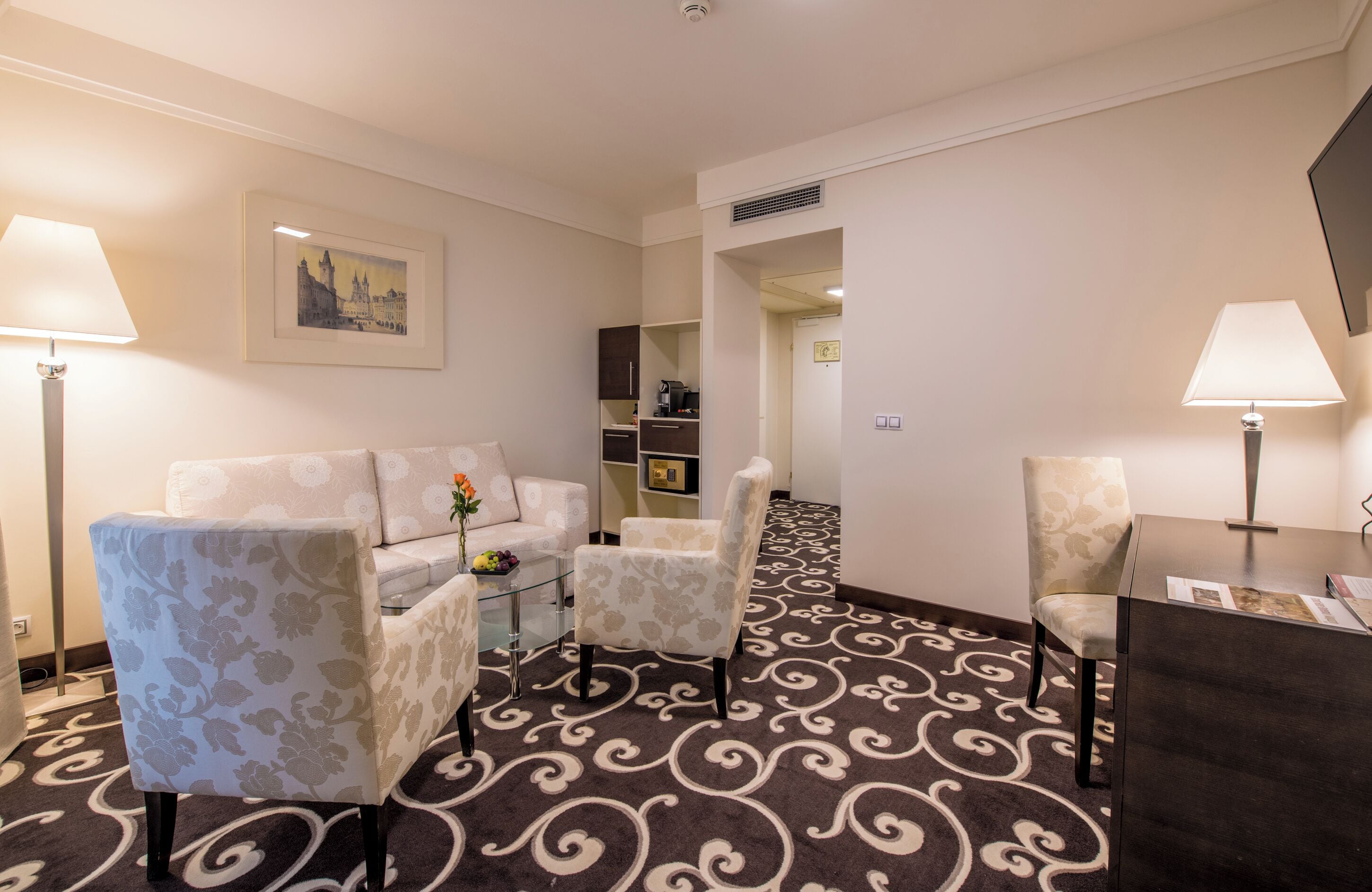 A hotel room with a sofa, two armchairs, a small table, a desk, and a patterned carpet.