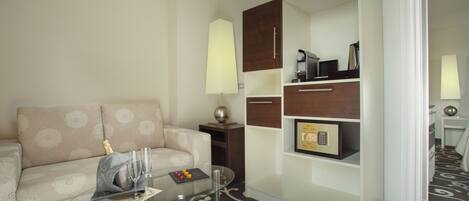 Una habitación de hotel moderna con una mesa de centro de vidrio, un sofá color beige, un armario de madera y una alfombra estampada.