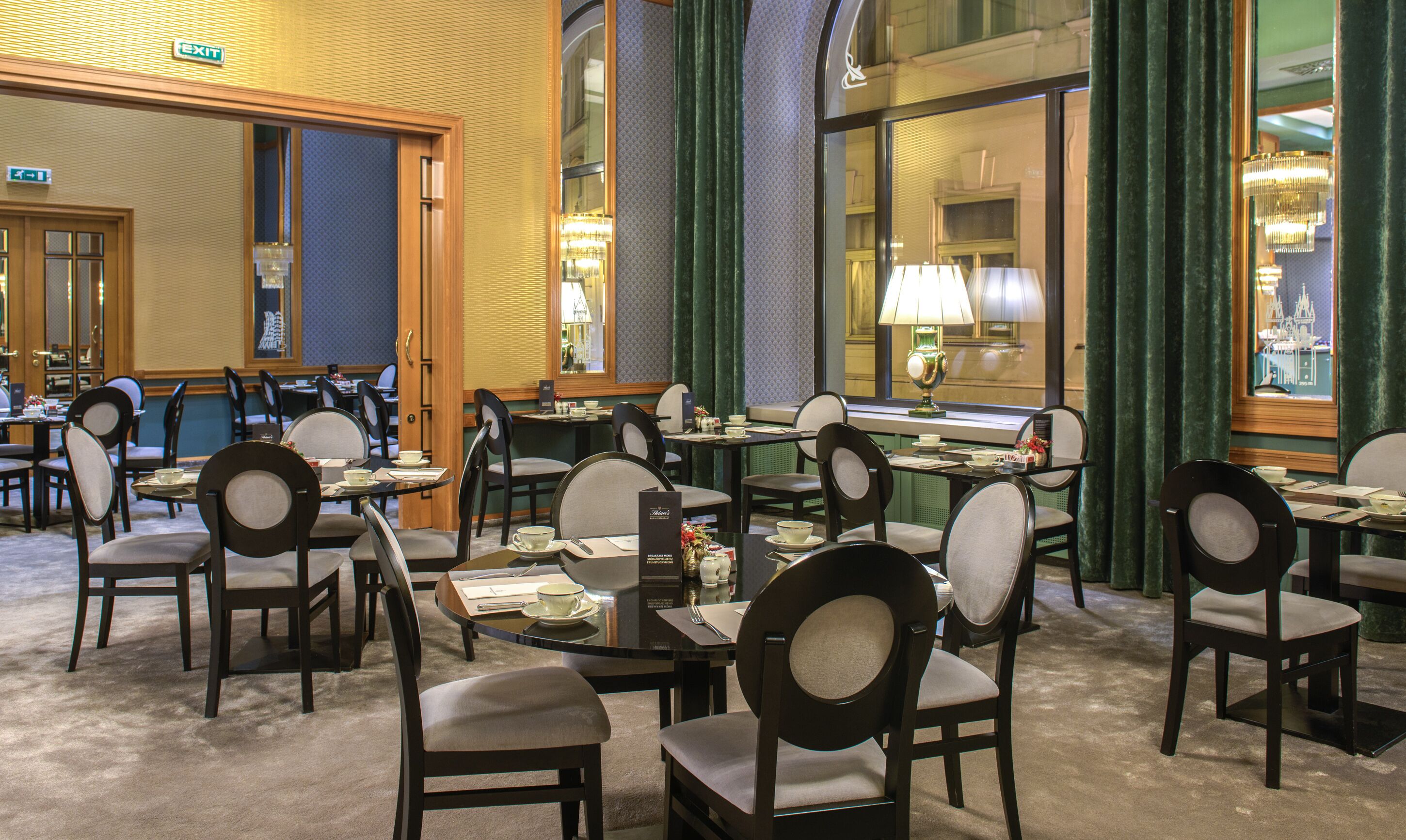 Un comedor de hotel con varias mesas y sillas, un candelabro y vistas a un edificio a través de amplios ventanales.