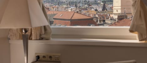 Una habitación con vista a un paisaje urbano histórico a través de una ventana, una lámpara en una mesita de noche y una silla en primer plano.