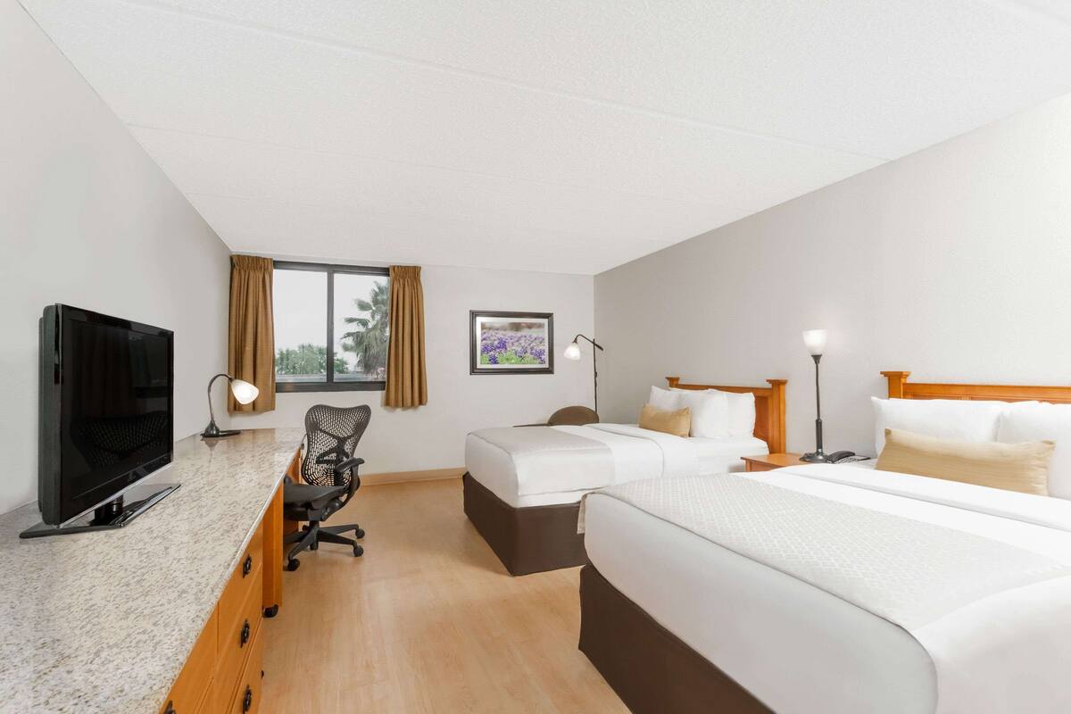 A hotel room with a TV, desk, two beds, a chair, and a window with curtains.
