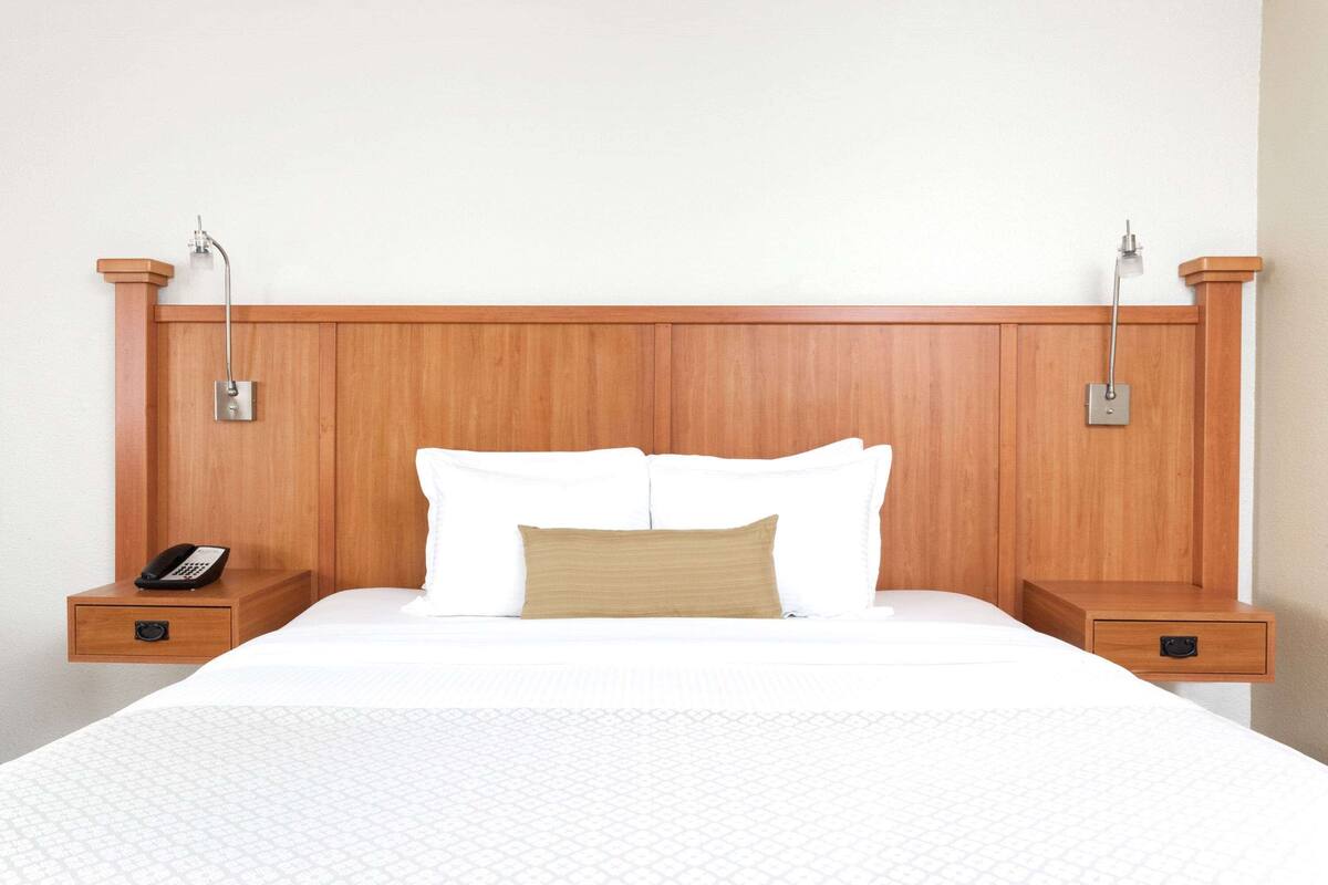 A hotel room with a wooden headboard, a white bed with a beige pillow, and a bedside table with a phone.