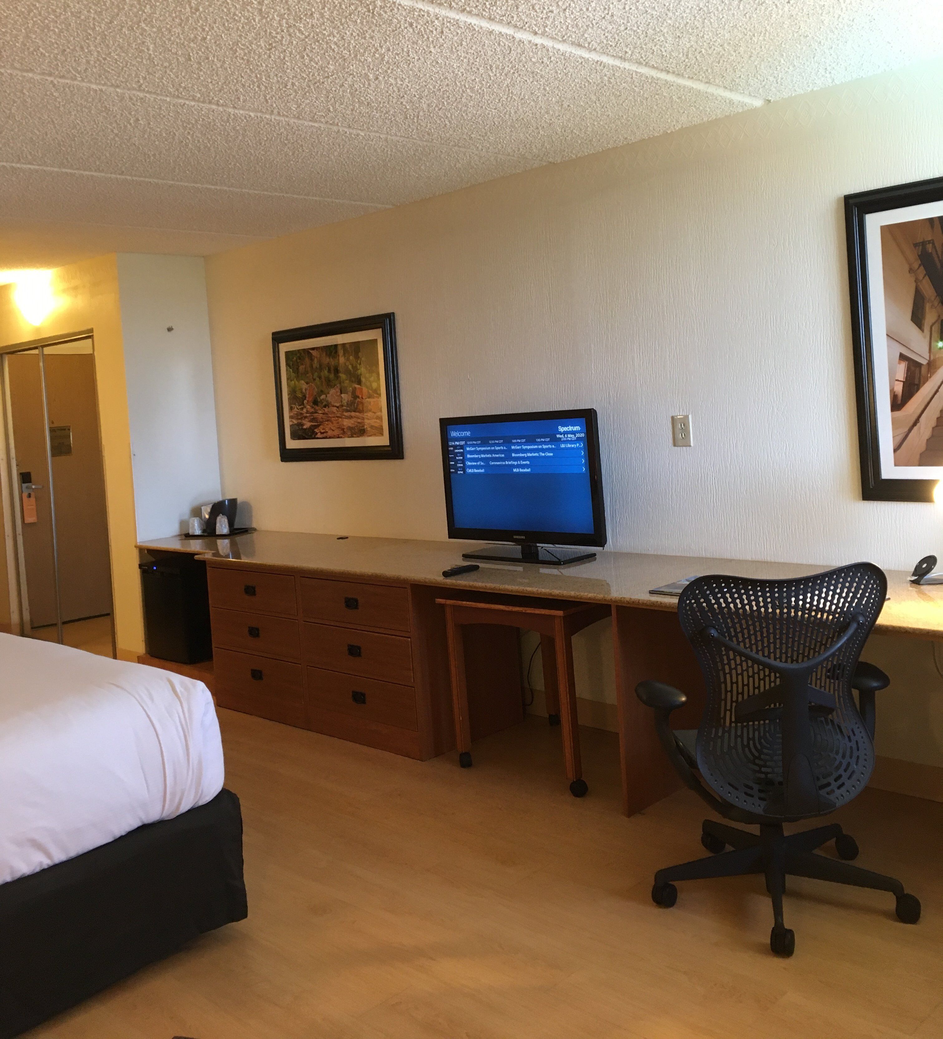 A hotel room with a bed, a desk with a computer, a chair, a framed picture, and a door.
