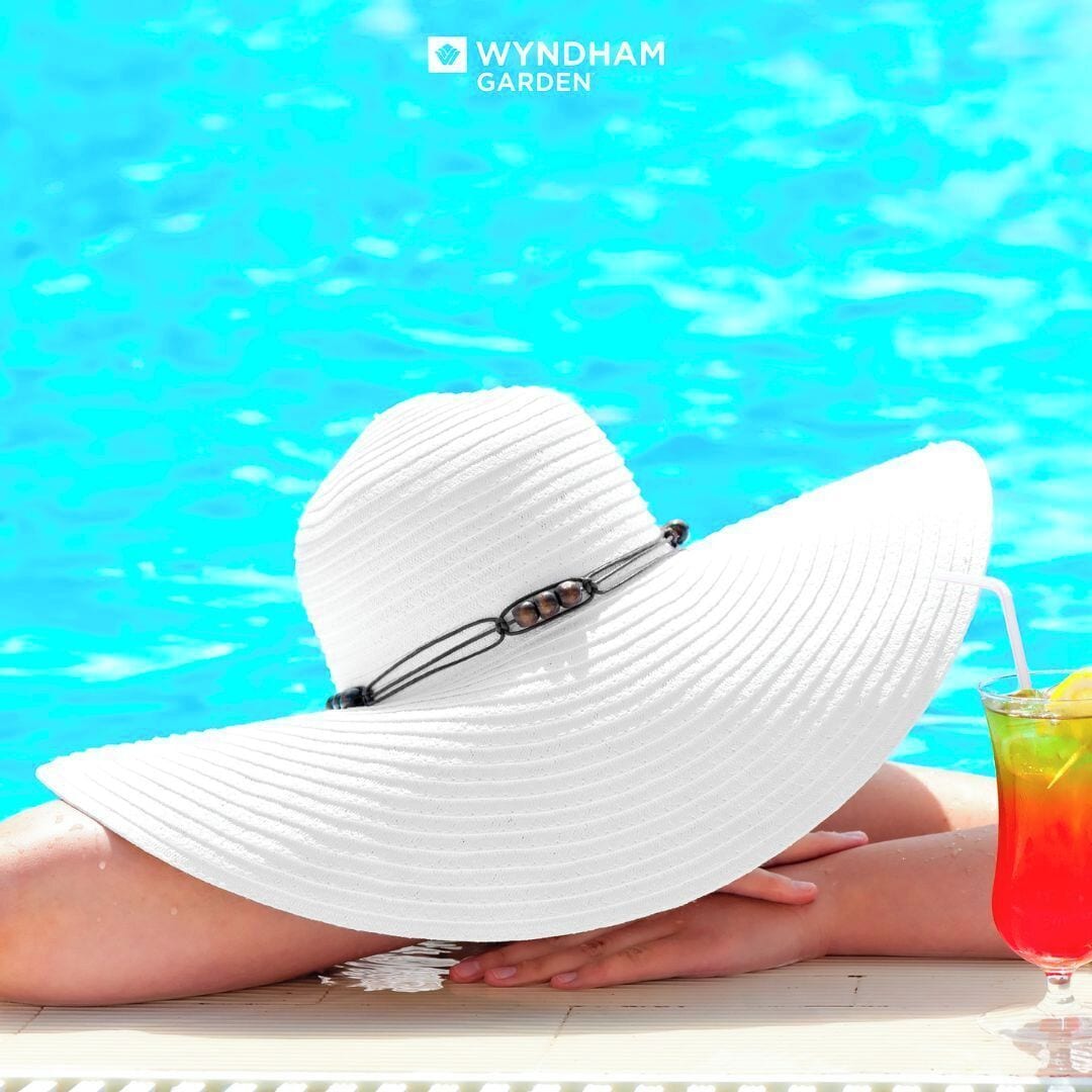 A person relaxing by a pool with a white sun hat and a drink.