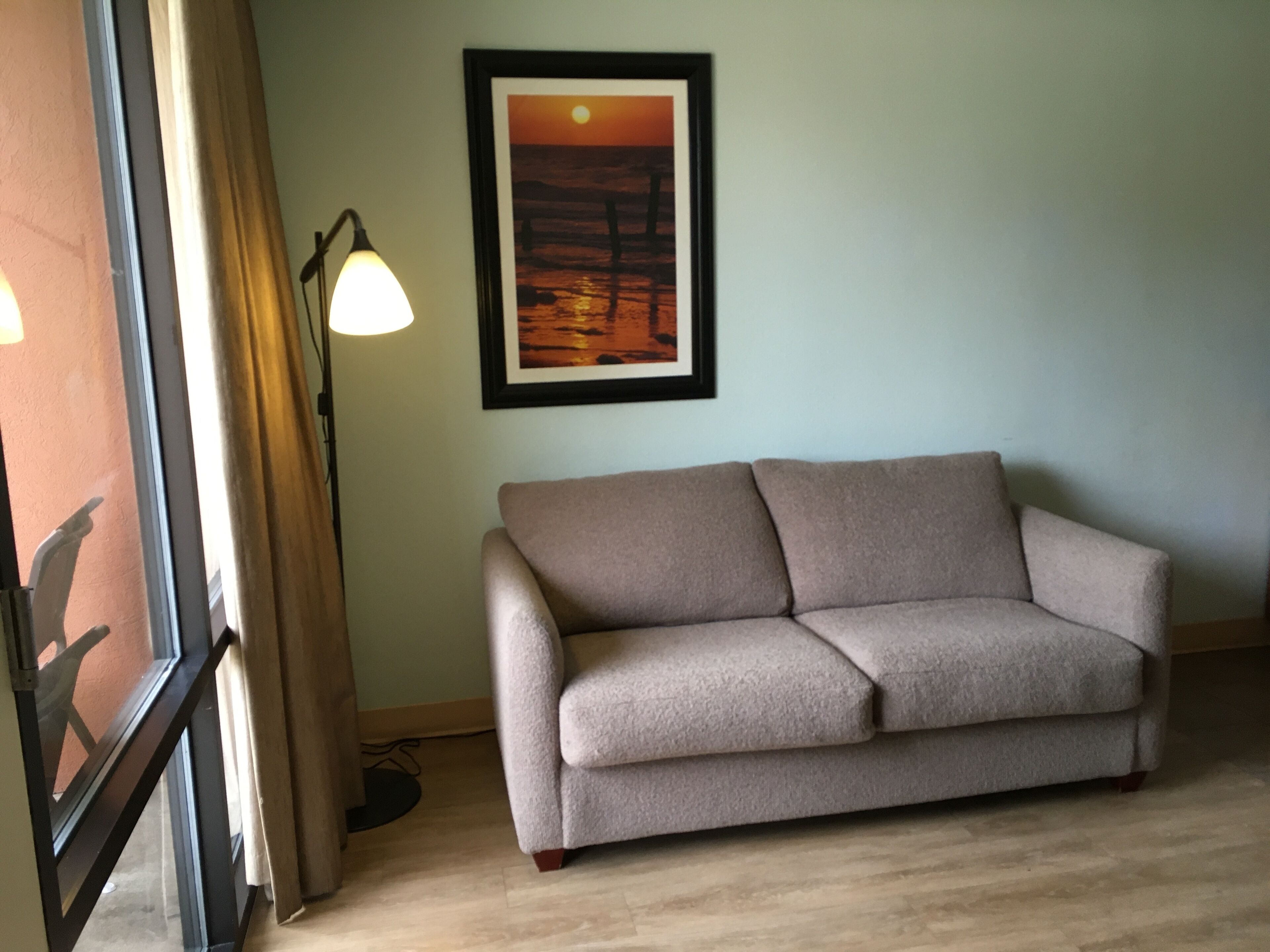 A room with a beige sofa, a framed sunset painting, a floor lamp, and a window with curtains.