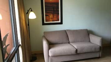 Et værelse med en beige sofa, et billede af en solnedgang i ramme, en gulvlampe og et vindue med gardiner.
