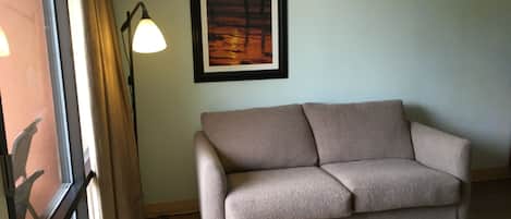 Quarto com sofá bege, pintura emoldurada de um pôr do sol, abajur e janela com cortinas.