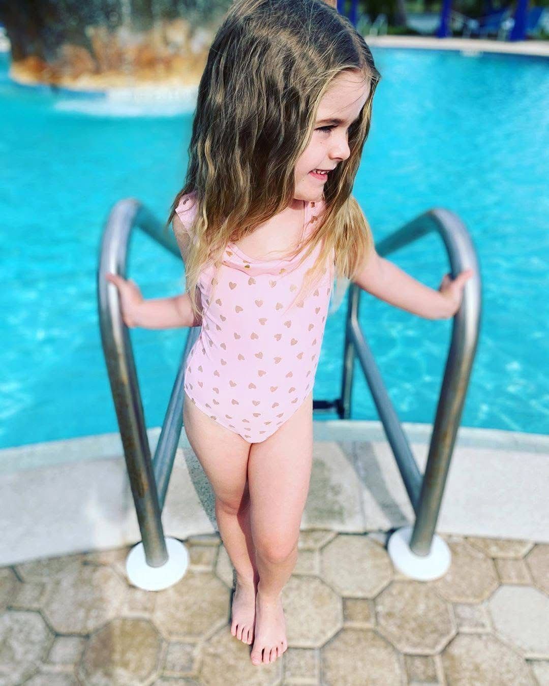 A child in a pink swimsuit with heart patterns standing by a pool with metal railings.