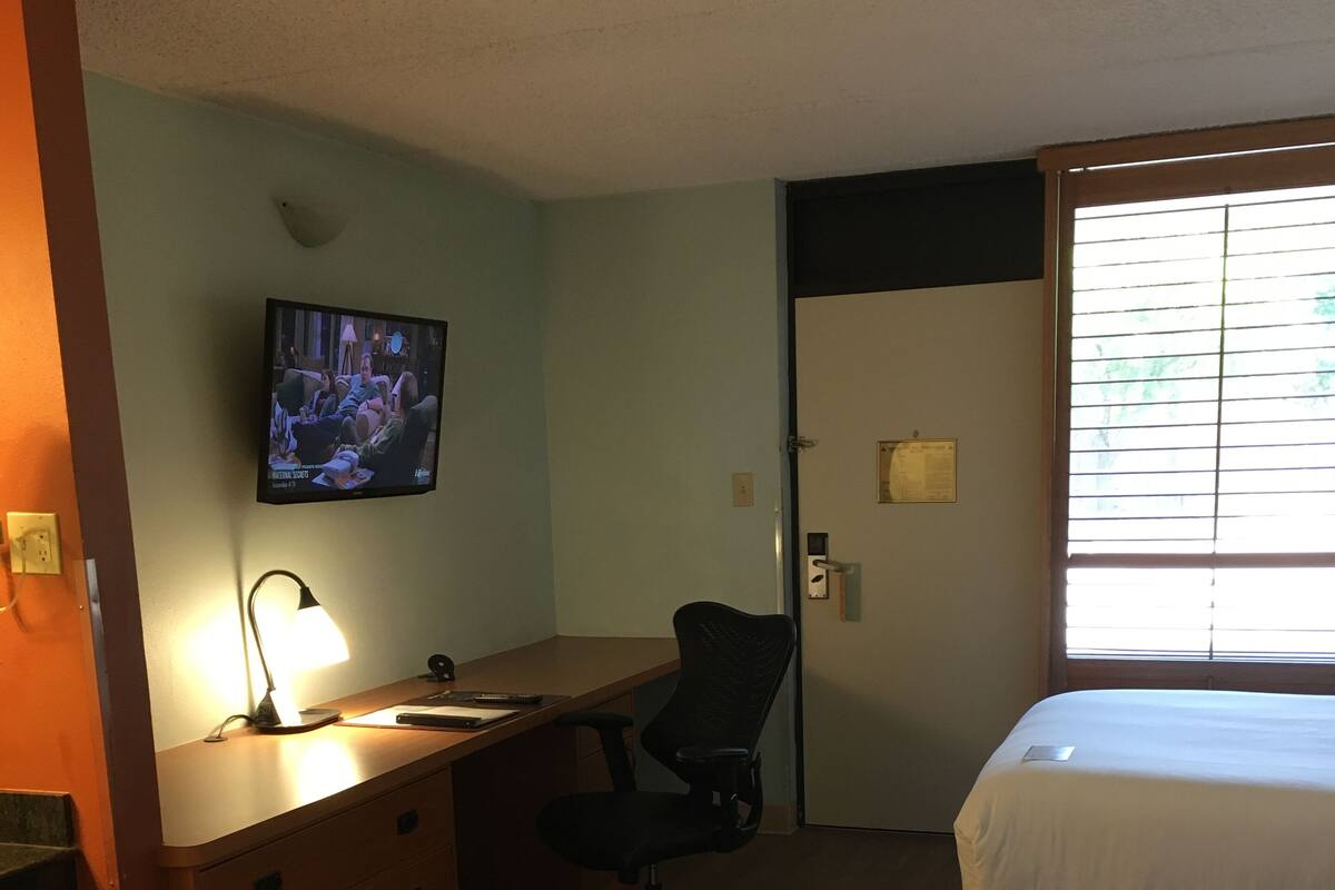 A hotel room with a wooden desk, a chair, a TV mounted on the wall, a bed, and a window with blinds.