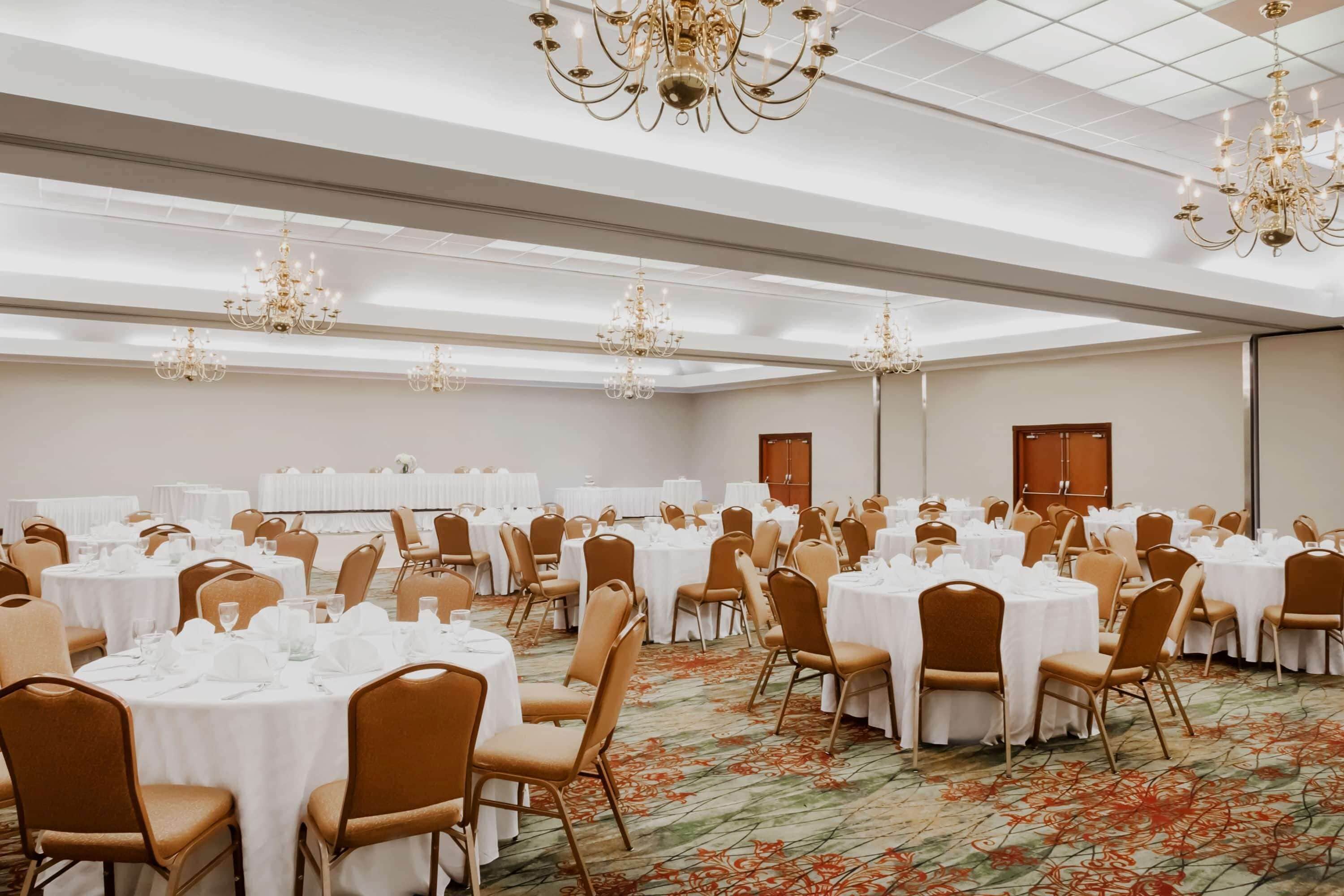 A banquet hall with round tables, chairs, chandeliers, and a carpeted floor.