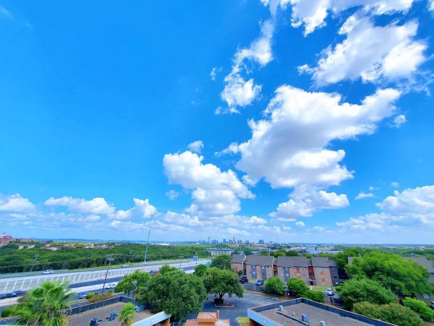 A view of a residential area with a clear blue sky and scattered clouds. There are multiple buildings, including a large brick structure and several smaller houses. A highway with moving vehicles is visible in the foreground.